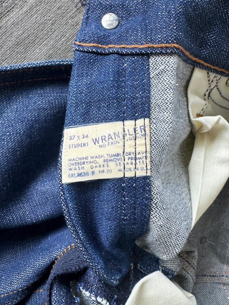Op de afbeelding: Close-up van een blauwe spijkerbroek met een zichtbaar label. Het label luidt "WRANGLER" en bevat wasvoorschriften en maat 27 x 34. De jeans is gemaakt in de VS.