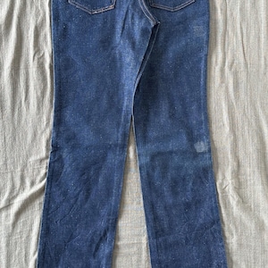 Op de afbeelding: Donkerblauwe denim jeans met een klassieke rechte pijp. Kenmerken zijn koperkleurige stiksels, een bruine leren patch met het "Wrangler" logo en traditionele vijf-zakken styling. De jeans liggen plat op een neutrale ondergrond.