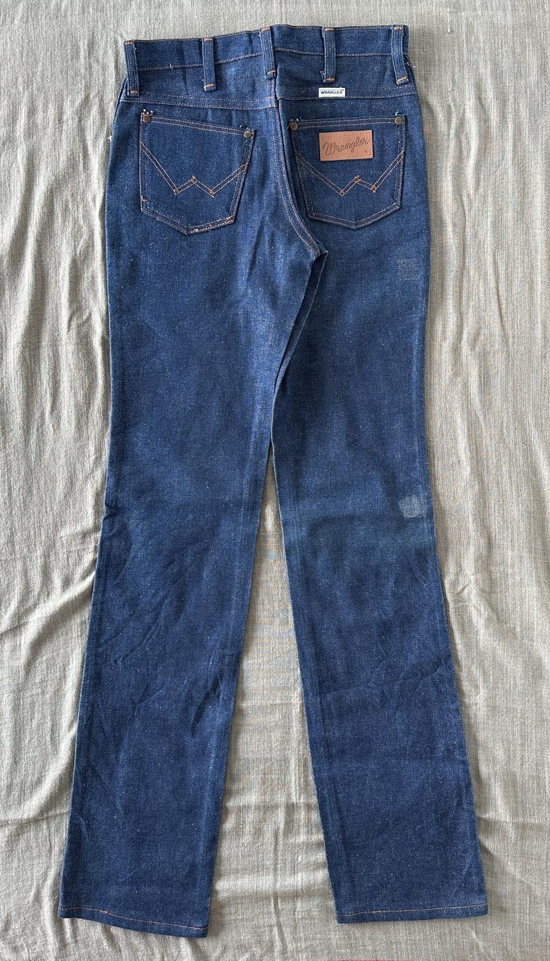 Op de afbeelding: Donkerblauwe denim jeans met een klassieke rechte pijp. Kenmerken zijn koperkleurige stiksels, een bruine leren patch met het "Wrangler" logo en traditionele vijf-zakken styling. De jeans liggen plat op een neutrale ondergrond.
