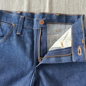 Op de afbeelding: Close-up van een blauwe denim jeans met een gedeeltelijk geopende ritssluiting. De jeans heeft een knoopsluiting en zichtbare stiksels in een contrasterende kleur. De details van de zak en de tailleband zijn duidelijk zichtbaar.