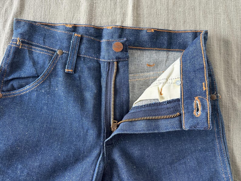 Op de afbeelding: Close-up van een blauwe denim jeans met een gedeeltelijk geopende ritssluiting. De jeans heeft een knoopsluiting en zichtbare stiksels in een contrasterende kleur. De details van de zak en de tailleband zijn duidelijk zichtbaar.