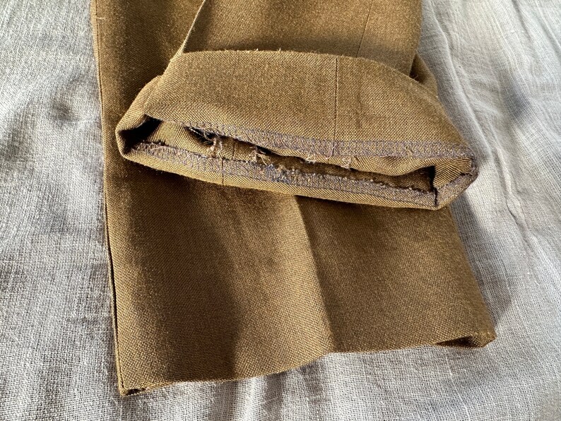 May include: Close-up of the hem of a pair of brown pants. The hem is folded over and sewn with a dark thread. The fabric is a woven twill.