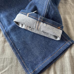 Op de afbeelding: Close-up van een donkerblauwe denim jeans met een omgeslagen zoom. De jeans heeft zichtbare oranje stiksels langs de naden en een lichtgrijze binnenkant op de zoom. De stof heeft een getextureerde uitstraling.