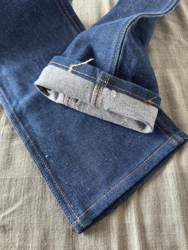 Op de afbeelding: Close-up van een donkerblauwe denim jeans met een omgeslagen zoom. De jeans heeft zichtbare oranje stiksels langs de naden en een lichtgrijze binnenkant op de zoom. De stof heeft een getextureerde uitstraling.
