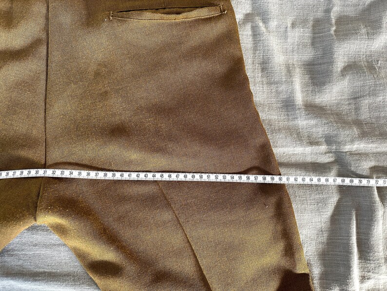 May include: Close-up of a pair of brown pants with a pocket on the back. A measuring tape is laid across the pants, showing a measurement of 70 inches.
