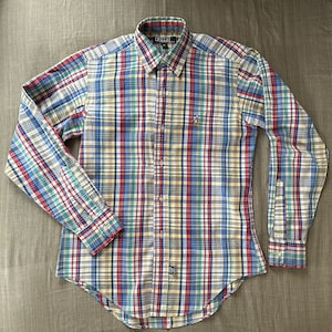 Puede incluir: Una camisa de manga larga a cuadros con botones y un pequeño logotipo bordado en el pecho izquierdo. La camisa es de cuadros multicolores con tonos de azul, rojo, verde y amarillo.