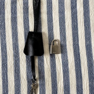 Puede incluir: Una clochette de cuero negro con una correa a juego y dos llaves, junto a un pequeño candado plateado. Los artículos están dispuestos sobre una tela a rayas azules y blancas. La clochette tiene una forma distintiva.
