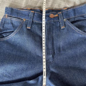 Op de afbeelding: Close-up van een donkerblauwe spijkerbroek met oranje stiksels. Een meetlint toont een lengte van ongeveer 94 cm. De jeans heeft een knoopsluiting en een klassiek zakontwerp.