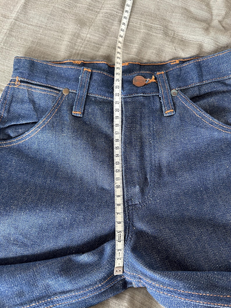 Op de afbeelding: Close-up van een donkerblauwe spijkerbroek met oranje stiksels. Een meetlint toont een lengte van ongeveer 94 cm. De jeans heeft een knoopsluiting en een klassiek zakontwerp.