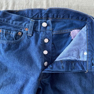 May include: A pair of blue denim jeans with a button fly and a single front pocket. The jeans are unbuttoned and the inside of the waistband is visible.