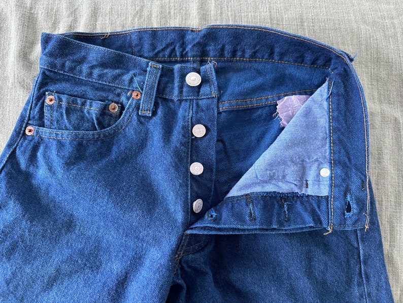 May include: A pair of blue denim jeans with a button fly and a single front pocket. The jeans are unbuttoned and the inside of the waistband is visible.