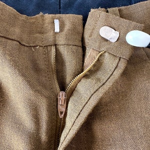 May include: Close-up of the front of a pair of brown linen pants with a zipper and a button closure. The zipper has a metal pull with the word "TALON" on it.
