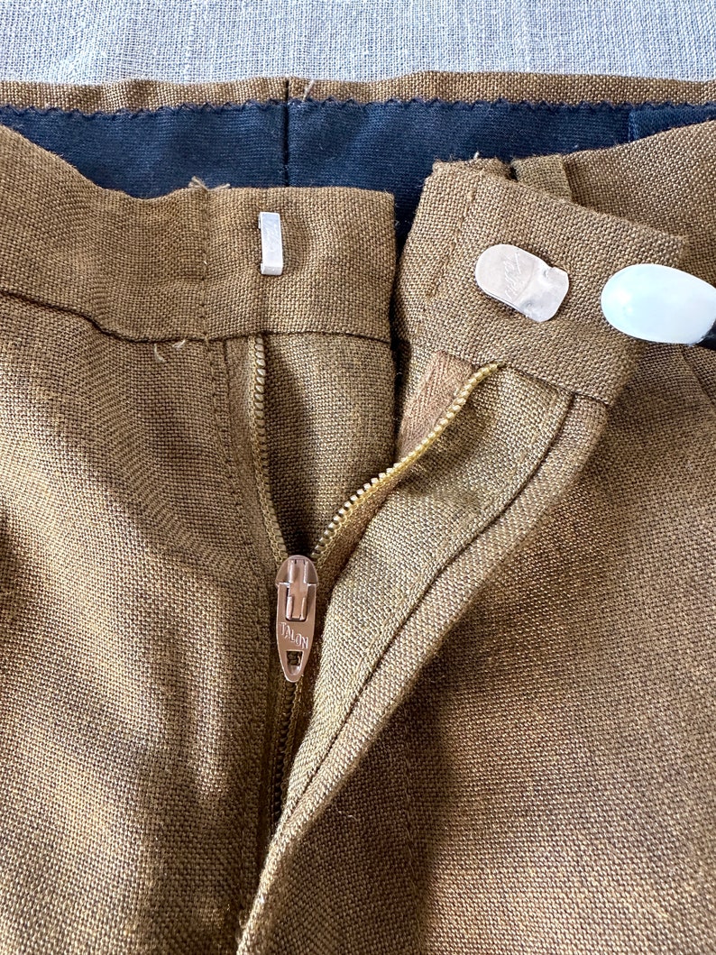 May include: Close-up of the front of a pair of brown linen pants with a zipper and a button closure. The zipper has a metal pull with the word "TALON" on it.