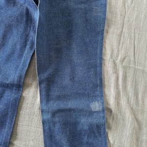 Op de afbeelding: Close-up van een blauwe spijkerbroek. De jeans heeft een klassiek ontwerp met vijf zakken met een vervaagde wassing en een kleine vlek op de broekspijp. De denim heeft een middellauw blauwe kleur.
