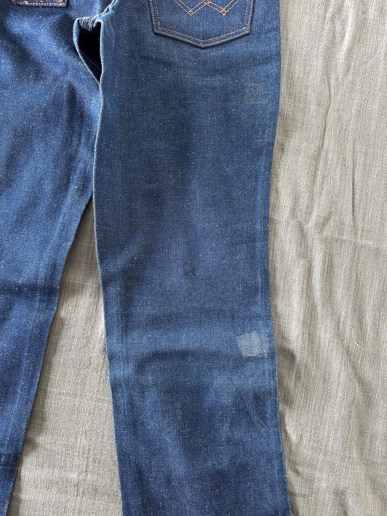 Op de afbeelding: Close-up van een blauwe spijkerbroek. De jeans heeft een klassiek ontwerp met vijf zakken met een vervaagde wassing en een kleine vlek op de broekspijp. De denim heeft een middellauw blauwe kleur.