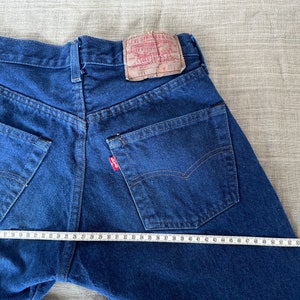 May include: A pair of blue denim jeans with a red Levi's label on the back pocket. The jeans are laid flat and measured with a tape measure, showing a waist measurement of 34 inches.