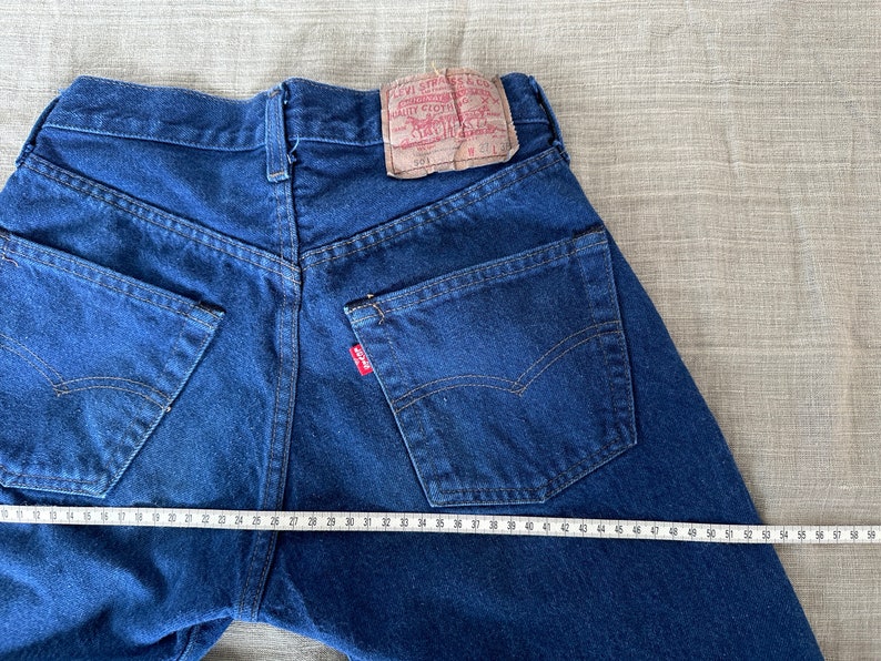 May include: A pair of blue denim jeans with a red Levi's label on the back pocket. The jeans are laid flat and measured with a tape measure, showing a waist measurement of 34 inches.