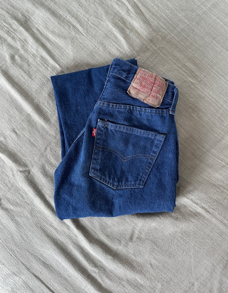 May include: A pair of blue denim jeans folded in half, with a brown Levi's label on the back pocket. The label has the text "Levi Strauss & Co.", "San Francisco, Cal.", "Original  Quality  Clothing", "W 31 L 32", and "501".