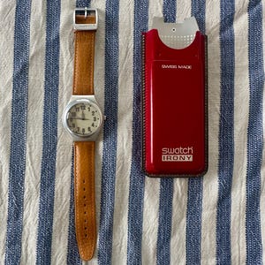 May include: A Swatch Irony wristwatch with a round silver-tone case, a cream-colored dial with black Arabic numerals, and a textured tan leather strap. Beside it is a red rectangular Swatch Irony protective case with the text SWISS MADE.