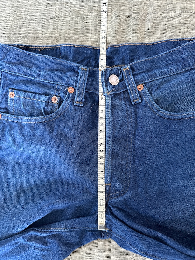 May include: A close-up of a pair of blue denim jeans with a measuring tape placed vertically along the center seam. The tape measures 35 inches.