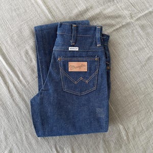 Op de afbeelding: Donkerblauwe denim jeans met het Wrangler-logo op de achterzak. De jeans is netjes opgevouwen en toont de achterzakken en riemlussen. De jeans is gemaakt van stevig denim materiaal.