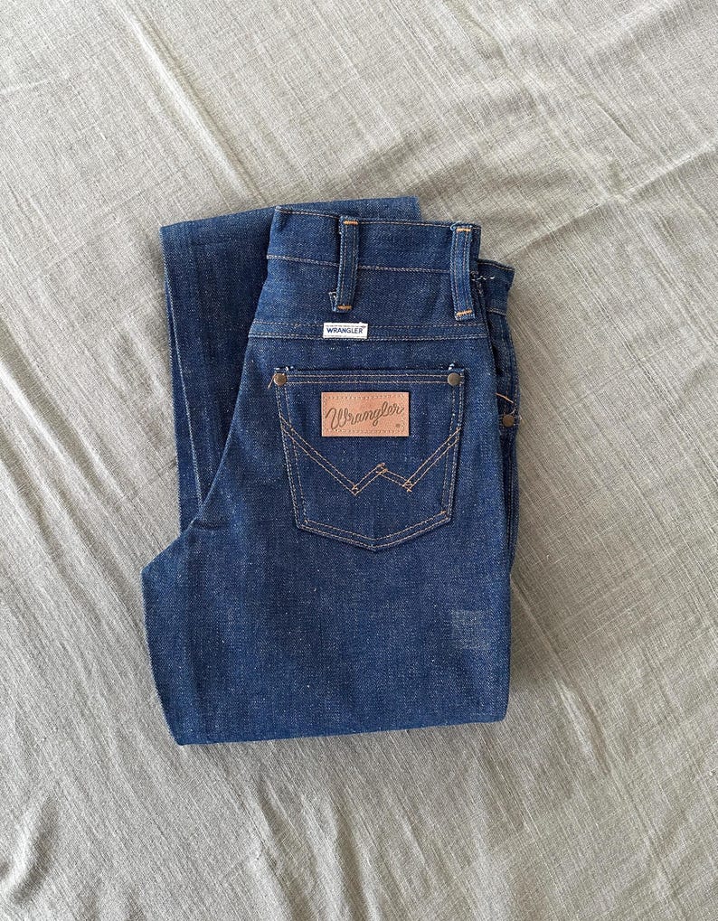 Op de afbeelding: Donkerblauwe denim jeans met het Wrangler-logo op de achterzak. De jeans is netjes opgevouwen en toont de achterzakken en riemlussen. De jeans is gemaakt van stevig denim materiaal.