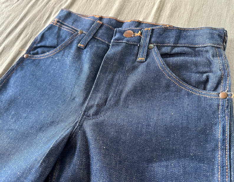 Op de afbeelding: Close-up van een donkerblauwe denim jeans. De jeans heeft een klassiek ontwerp met vijf zakken, koperkleurige klinknagels en stiksels. De stof heeft een licht versleten uiterlijk, wat wijst op een vintage of geliefd item.