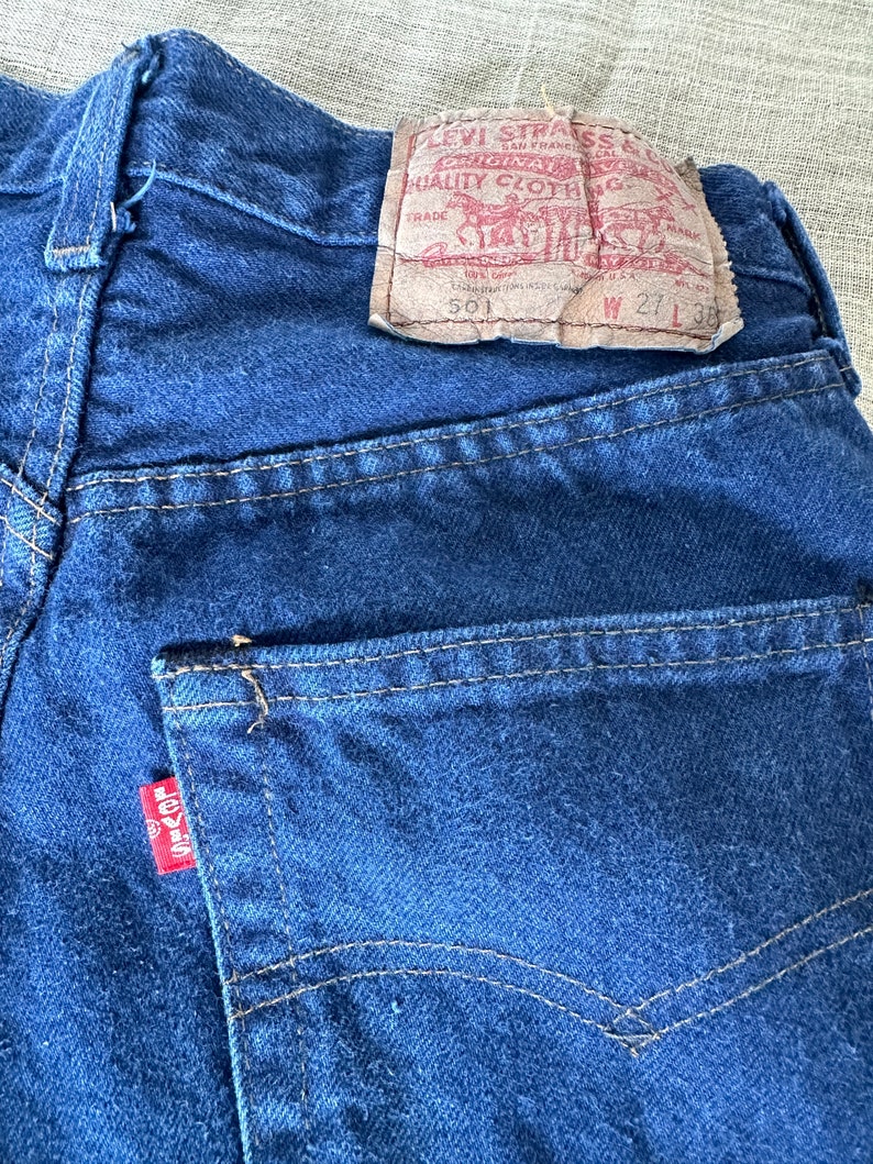 May include: A pair of blue denim jeans with a Levi's label on the back pocket. The label reads "Levi Strauss & Co. Original Quality Clothing" and has the size "W 27 L 32" printed on it.