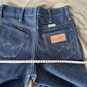 Op de afbeelding: Donkerblauwe Wrangler jeans van denim met klassieke stiksels op de achterzakken en een leren Wrangler patch. De jeans worden gemeten met een meetlint, met een taillemaat van ongeveer 61 cm.