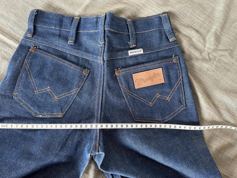 Op de afbeelding: Donkerblauwe Wrangler jeans van denim met klassieke stiksels op de achterzakken en een leren Wrangler patch. De jeans worden gemeten met een meetlint, met een taillemaat van ongeveer 61 cm.
