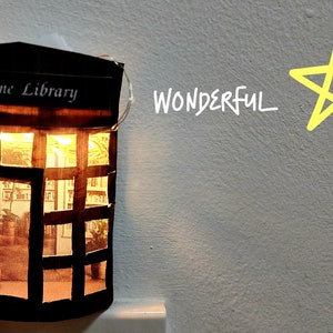 May include: A small cardboard model of a home library with a lit string of fairy lights inside. The model has a sign that says "Home Library" and a window with a view of bookshelves. The background is a light grey wall with the word "WONDERFUL" written in white and a yellow star drawn on the wall.