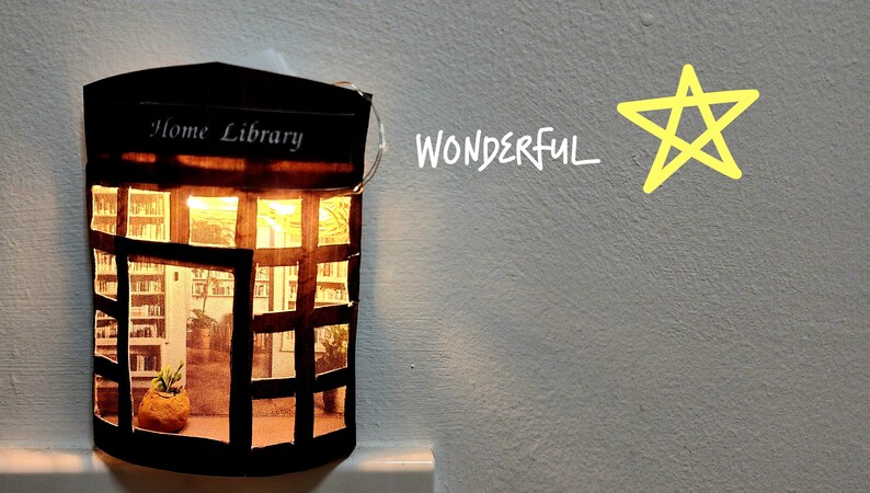 May include: A small cardboard model of a home library with a lit string of fairy lights inside. The model has a sign that says "Home Library" and a window with a view of bookshelves. The background is a light grey wall with the word "WONDERFUL" written in white and a yellow star drawn on the wall.