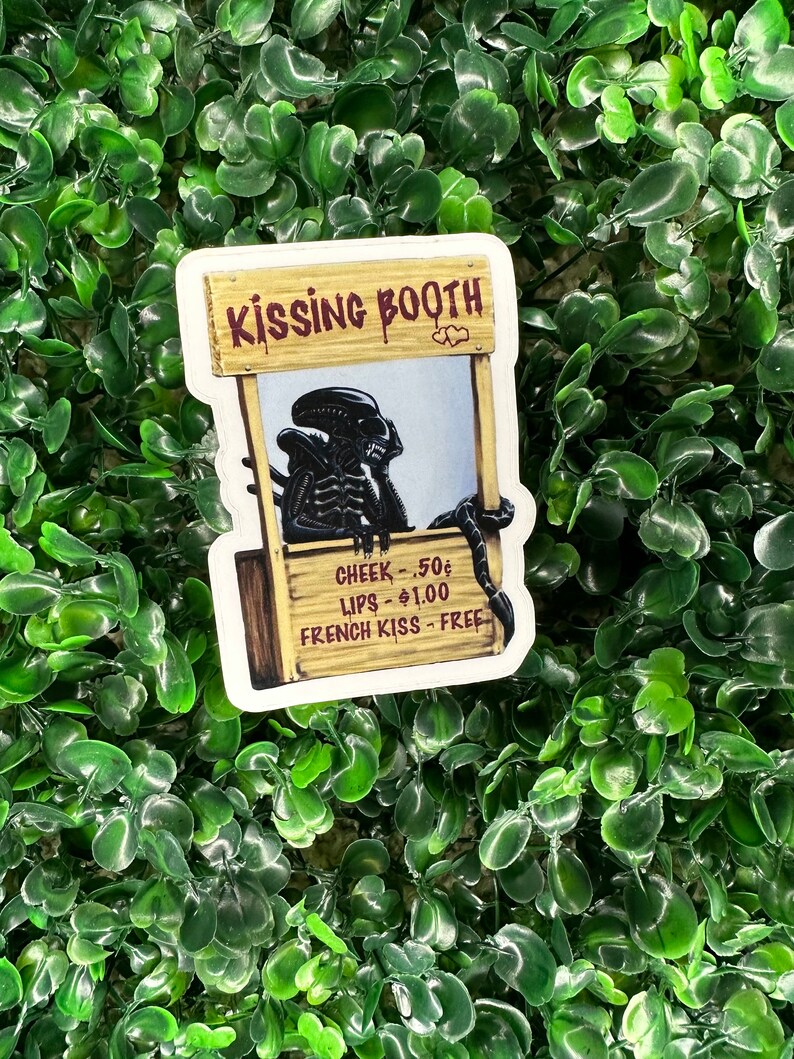 Puede incluir: Una pegatina que representa un puesto de besos con un personaje alien&iacute;gena. El letrero del puesto dice "KISSING BOOTH" en letras marrones. El alien&iacute;gena es negro con un dise&ntilde;o detallado. El puesto tiene una lista de precios: "CHEEK - 50&cent;, LIPS - $1.00, FRENCH KISS - FREE."