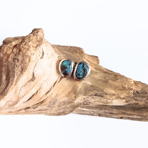 Può includere: Un paio di orecchini a lobo con pietre scure di forma irregolare e riflessi iridescenti blu e verdi, ciascuna incastonata in una montatura in metallo color argento liscio e lucido che segue la forma organica e asimmetrica della pietra.