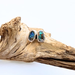 Op de afbeelding: Een paar oorknopjes met onregelmatige, gepolijste opaalstenen met levendige blauwe en groene flitsen, elk gezet in een eenvoudige zilverkleurige metalen zetting. De stenen hebben een donkere, aardse basiskleur met een hoogglans afwerking.