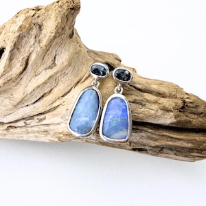 May include: A pair of silver earrings with blue and black gemstones. Each earring features a large, light blue, oval-shaped stone with a smaller, dark blue, oval-shaped stone above it. The earrings are displayed on a piece of driftwood.