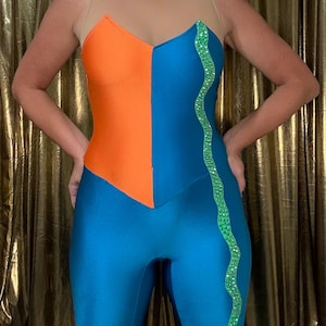 Peut inclure: Un justaucorps bicolore bleu et orange vif, avec un haut en résille nude transparent. Une bande ondulée de paillettes vertes orne le côté bleu. Le justaucorps est moulant, probablement pour la danse ou la performance.