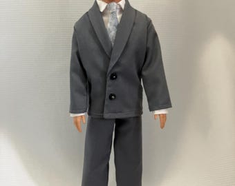 May include: A doll wearing a gray suit with a white collared shirt and a patterned tie. The suit has a two-button closure. The doll has blonde hair and is standing on a white surface.