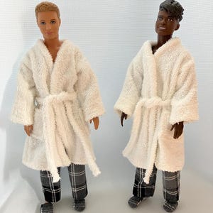May include: Two doll figures wearing matching white terry cloth robes with tie belts and plaid pajama pants. The dolls have different skin tones and hairstyles. The robes have a soft, fluffy texture, and the pajama pants are black, white, and gray.