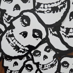 May include: A collection of white and black skull-shaped stickers. Each sticker features a stylized skull design with black outlines and details. The stickers are arranged in a close-up, overlapping pattern.