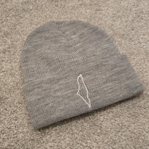 May include: A gray beanie hat with a white embroidered outline of a country. The hat is made of a soft, knitted material and is laying on a light gray carpet.