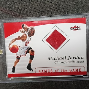 May include: A Michael Jordan Chicago Bulls-guard trading card in a protective case. The card features an image of Jordan in action, a red jersey swatch, and the text "NAMES of the GAME."
