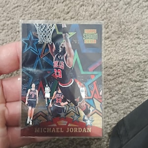 Può includere: Una carta di basket di Michael Jordan della serie Topps Stadium Club. La carta mostra Jordan in una schiacciata, con la sua maglia dei Chicago Bulls con il numero 23. Lo sfondo presenta stelle e forme geometriche.
