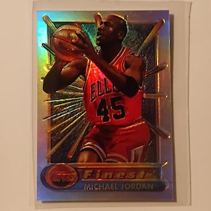 Può includere: Una carta di basket di Michael Jordan con una foto del giocatore in una divisa rossa dei Chicago Bulls, che tiene un pallone da basket. La carta ha uno sfondo olografico argentato con la scritta "Finest" e "Michael Jordan".