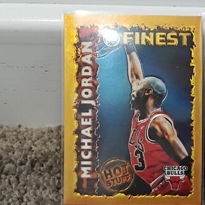 May include: A Michael Jordan Finest basketball trading card featuring a gold border with a flame-like edge. The card shows Jordan in a red Chicago Bulls jersey reaching for a basketball hoop, with the text "HOT STUFF" and "FINEST" printed on it.