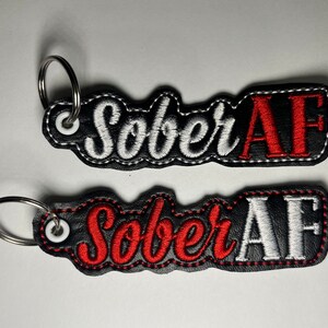 May include: Two black leather keychains with a white and red embroidered design that says "Sober AF".