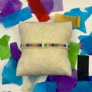 May include: A colourful beaded bracelet with a gold accent on a white cushion. The bracelet is made of small, colourful beads and has a gold accent in the centre. The bracelet is on a white cushion, which is surrounded by colourful tissue paper.
