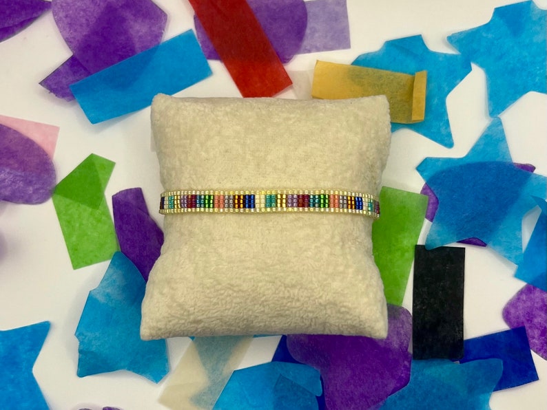 May include: A colourful beaded bracelet with a gold accent on a white cushion. The bracelet is made of small, colourful beads and has a gold accent in the centre. The bracelet is on a white cushion, which is surrounded by colourful tissue paper.