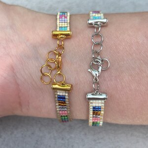 May include: Two beaded bracelets with colourful stripes and gold and silver clasps. The bracelets are on a person's wrist.