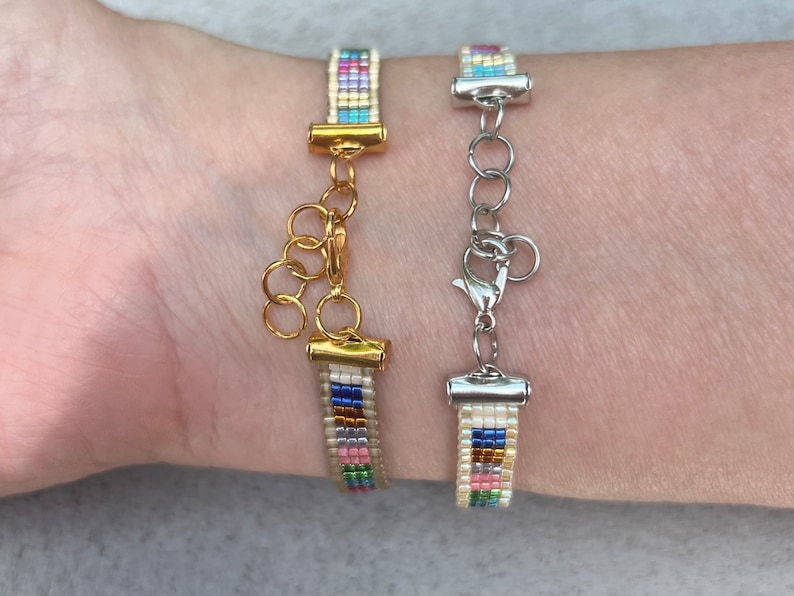 May include: Two beaded bracelets with colourful stripes and gold and silver clasps. The bracelets are on a person's wrist.